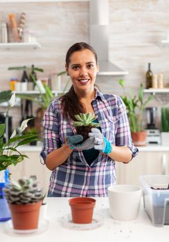 Portrait of happy woman holding succulent plant sitting on the table in kitchen. Woman replanting flowers in ceramic pot using shovel, gloves, fertil soil and flowers for house decoration.