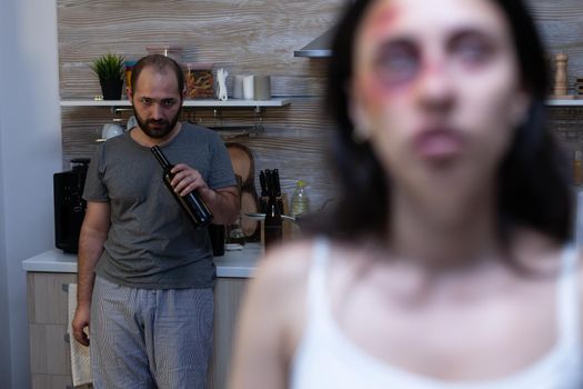 Violent man drinking alcohol from bottle having addiction while abused hurt woman sitting crying with bruises on face. Terrified wife being victim of domestic violence and alcoholism aggression