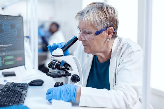 Senior chemist scientist doing genetic investigation using microscope. Elderly researcher carrying out scientific research in a sterile lab using a modern technology.