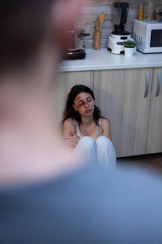 Domestic violence victim sitting on floor because of social problems. Abusive husband beating terrified helpless wife with bruises on face. Woman with physical trauma, aggression