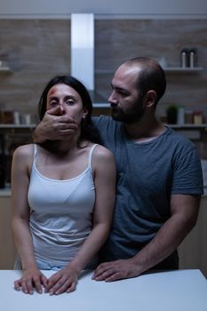 Aggresive husband being cruel to bruised wife at home. Victim woman dealing with domestic violence and injury from partner. Caucasian man holding hand on mouth harassing terrified person