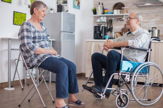 Old woman and her disabled husband in wheelchair chatting in kitchen.Elderly person having a conversation with husband in kitchen. Living with disabled person with walking disabilities