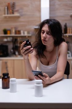 Sick woman with migraine looking at container with medicine and smartphone for prescription. Person with headache holding bottle of drugs, medication, remedy, medicament, treatment