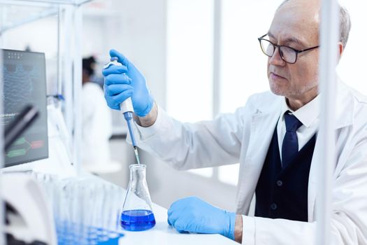 Experienced scientist working in biotechnological laboratory with sterile dropper and blue liquid. Senior professional chemist using pippete with blue solution for microbiology tests.