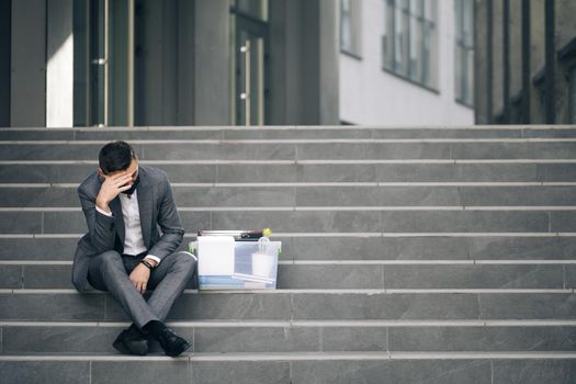 Fired caucasian male office worker in medical mask sitting on stairs in depression with box of stuff. Anxious concept. Workless man in despair. Unemployed businessman lost his business.