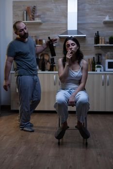 Depressed wife dealing with physical trauma and domestic violence smoking cigarette while listening to aggressive violent husband holding bottle of alcohol. Couple with social problems