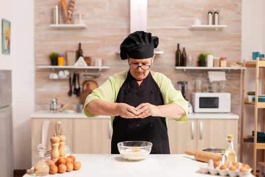 Cracking eggs into wheat flour following traditional recipe in home kitchen. Elderly pastry chef cracking egg on glass bowl for cake recipe in kitchen, mixing by hand, kneading.