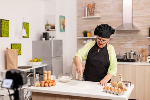 Senior woman creating content for culinary blog preparing tasty bread. Retired blogger chef influencer using internet technology communicating, shooting blogging on social media with digital equipment.