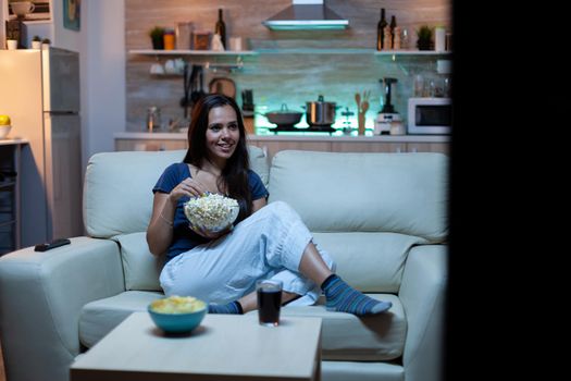 Amused lady watching Tv show in the night sitting on couch. Young happy excited home alone woman enjoying the evening sitting on comfortable sofa dressed in pajamas eating popcorn in front television.