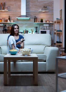 Young cheerful lady reasting watching TV sitting on comfortable couch at home. Amused woman enjoying the evening sitting on cozy sofa dressed in pajamas eating popcorn in front television.