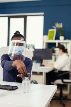 African woman in corporation office applying hand sanitizer wearing face mask during coronavirus. Businesswoman in new normal workplace disinfecting while colleagues working in background.