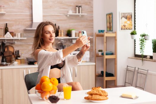 Sensual woman taking photos using smartphone in home kitchen while wearing sexy underwear. Seductive woman with tattoos using smartphone wearing temping underwear in the morning.