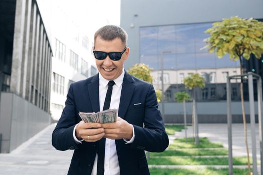 Rich man in stylish suit wearing sunglasses counting money and walking near office building. Looking successful, confident.