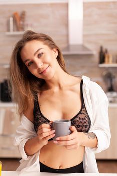 Sexy blonde lady in lingerie drinking coffee during breakfast enjoying the morning. Young attractive woman with tattoos in seductive underwear holding cup of tea relaxing in the kitchen smiling.