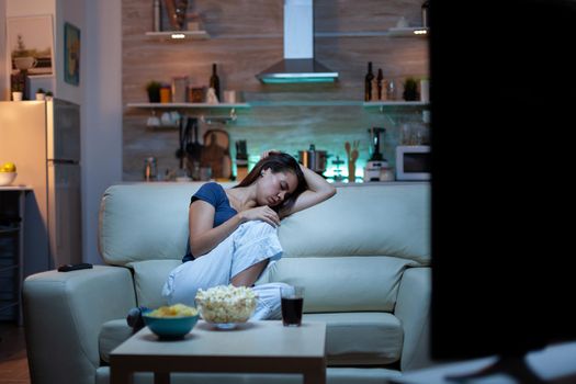 Young lady sleeping on couch watching tv. Tired exhausted lonely sleepy woman in pajamas falling asleep on sofa in front television, closing eyes while watching movie at night in living room