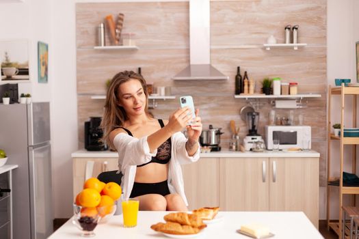 Seductive woman in sexy lingerie taking selfie using smartphone in home kitchen. Attractive lady with tattoos using smartphone wearing temping underwear in the morning.