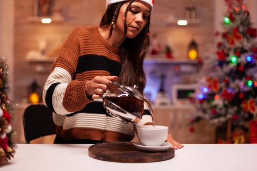 Woman with winter clothes pouring cup of tea from kettle at home decorated for christmas eve festivity. Caucasian person in festive kitchen enjoying seasonal drink feeling cozy indoors