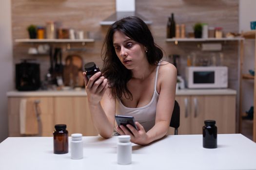 Portrait of woman with painful migraine holding bottle of medicine reading prescription for pills, drugs, remedy, medication, treatment and medicament on smartphone against headache