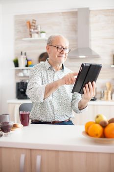 Happy old man in kitchen during breakfat using tablet pc. Elderly person with tablet portable pad PC in retirement age using mobile apps, modern internet online information technology with touchscreen