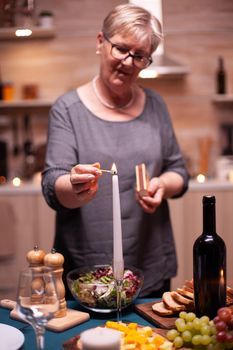 Candle burning on table for romantic dinner of senior couple in kitchen. Elderly woman waiting her husband for a romantic dinner. Mature wife preparing festive meal for anniversary celebration.