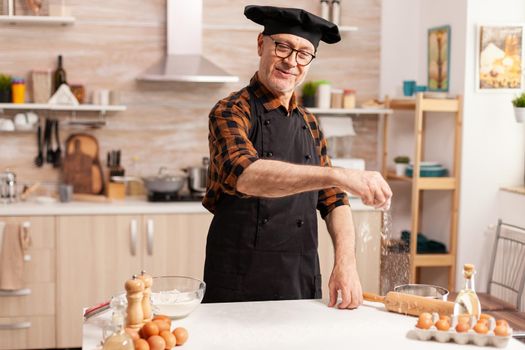 Senior man wearing apron smiling while sprinkling what flour with hand on kitchen table. Retired chef with bonete and apron, in kitchen uniform sprinkling sieving sifting ingredients by hand.