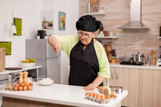 Baker spreading flour on wooden table at home in modern kitchen. Happy elderly chef with uniform sprinkling, sieving sifting raw ingredients by hand baking homemade pizza