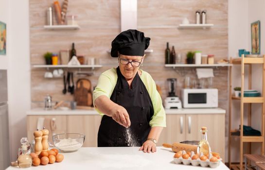 Chef sprinkling baking powder on home kitchen table wearing bonete and apron. Happy elderly chef with uniform sprinkling, sieving sifting raw ingredients by hand baking homemade pizza