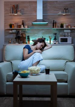 Tired woman closing eyes while watching movie at night. Tired exhausted lonely sleepy housewife in pajamas sleeping in front of television sitting on cozy couch in living room at home.