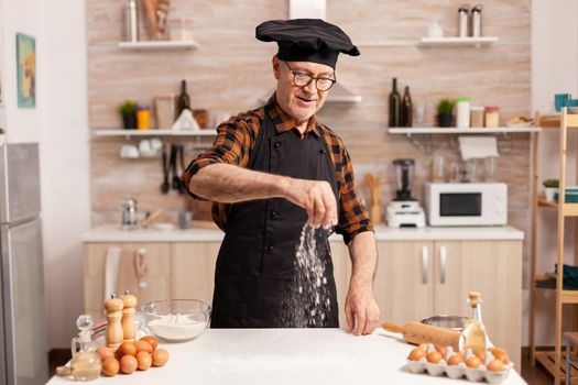 Senior baker sprankling wheat flour to prepare delicious cookies wearing bonete. Retired chef with bonete and apron, in kitchen uniform sprinkling sieving sifting ingredients by hand.