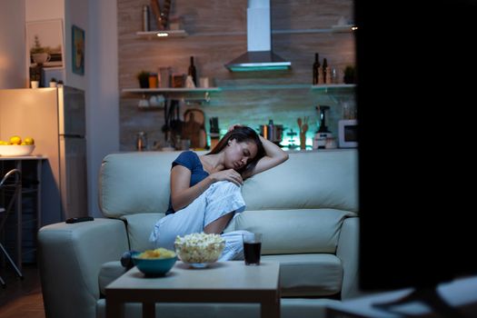 Woman sleeping on a sofa in front of TV while watching a bored movie. Tired exhausted lonely sleepy lady in pajamas falling asleep sitting on cozy couch in living room, closing eyes at night