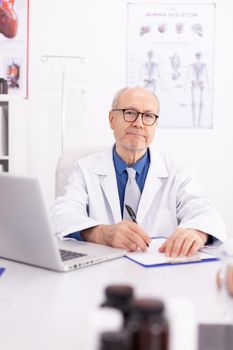 Mature doctor in hospital office wearing lab coat while looking at camera. Medical practitioner using notebook in clinic workplace , confident, expertise, medicine.