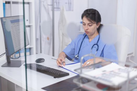 Medical staff writing notes on clipboard sitting at desk in hospital office. Healthcare physician specialist in medicine providing health care services treatment examination.