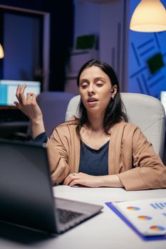 Businesswoman using earbuds in the course of online call doing overtime. Woman working on finance during a video conference with coworkers at night hours in the office.