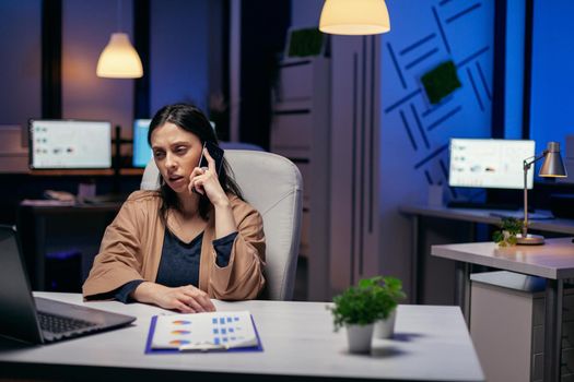 Worried businesswoman talking with clients in the course of smartphone conversation. Woman entrepreneur working late at night in corporate business doing overtime in the course of phone call.