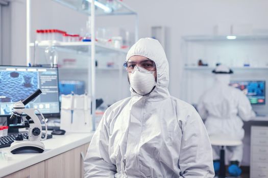 Chemist feeling tired from work for a long time in laboratory sitting at workplace. Overworked researcher dressed in protective suit against invection with coronavirus during global epidemic.