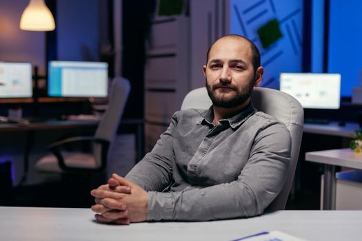 Tired serious freelancer looking at camera holding hands together. Smart businessman sitting at his workplace in the course of late night hours doing his job.