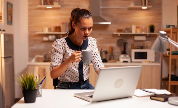 Tired woman holding cup of coffee in kitchen late at night while working on laptop. Employee using modern technology at midnight doing overtime for job, business, busy, career, network, lifestyle.