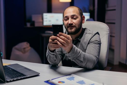 Caucasian freelancer messaging in the course of evening sitting at desk. Businessman using his cell to text message while working late at night in the office to finish a deadline.