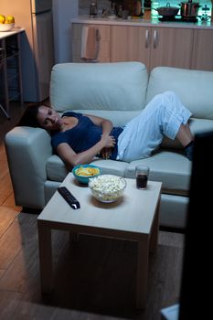 Young woman in pijamas resting lying on couch in front tv smiling. Happy, amused, lonely lady enjoying the evening sitting on comfortable sofa watching television eating popcorn and snacks