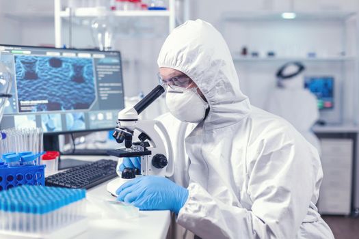 Doctor researching and evaluating virus using microscope in laboratory wearing ppe. Scientist in protective suit sitting at workplace using modern medical technology during global epidemic.