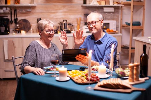 Mature man and woman waving at tablet pc during video conference in kitchen celebrating relationship. Couple sitting at the table, browsing, talking, using internet, celebrating their anniversary in dining room.