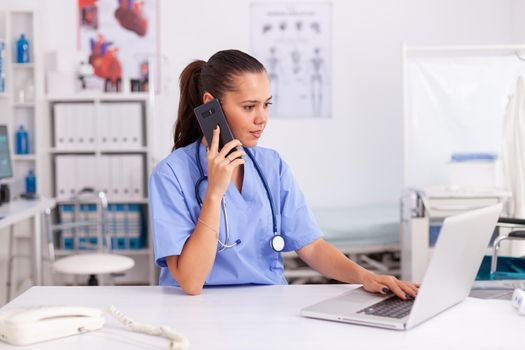 Medical practitioner using telephone and laptop in hospital office wearing blue uniform. Health care physician sitting at desk using computer in modern clinic looking at monitor.