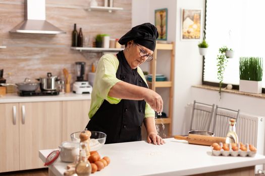 Elderly woman wearing apron while sprinkling flour in kitchen table. Happy elderly chef with uniform sprinkling, sieving sifting raw ingredients by hand baking homemade pizza.