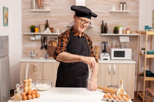 Smiling experienced male baker making homemade cookies wearing apron and bonete. Retired senior chef in kitchen uniform sprinkling sieving sifting ingredients by hand.