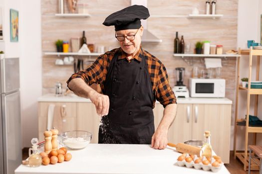 Senior chef preparing homemade bread sprinkling wheat flour on kitchen table. Retired senior chef with bonete and apron, in kitchen uniform sprinkling sieving sifting ingredients by hand.