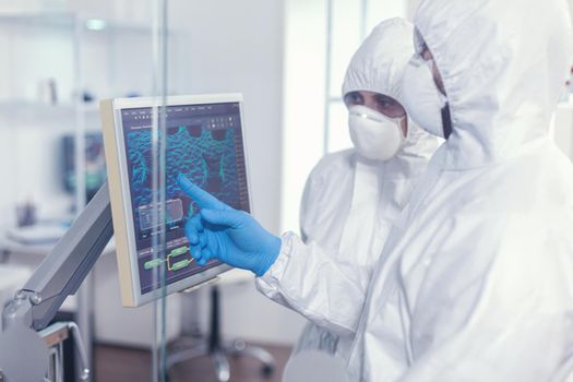 Chemists in protection suit discussing about changes in vaccine composition in equipped lab pointing desktop. Doctors examining virus evolution using high tech researching diagnosis against covid19