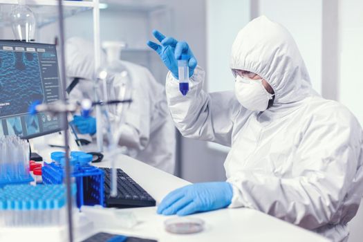 Scientist in lab coversall looking closely at sample in test tube in the course of coroanvirus. Researcher in laboratory wearing coverall doing research and analyzing substance during global pandemic with covid19.