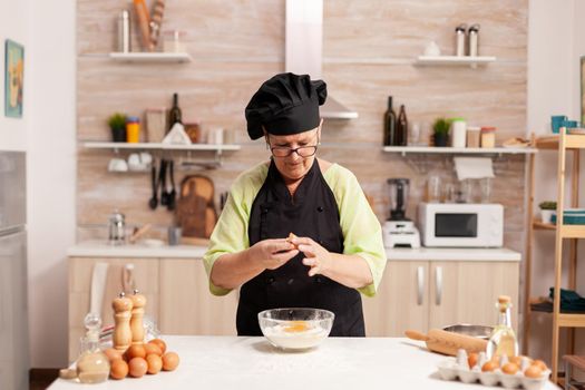 Chef hands with flour and eggs in preparation process for baking. Elderly pastry chef cracking egg on glass bowl for cake recipe in kitchen, mixing by hand, kneading.