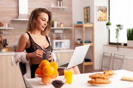 Sexy woman relaxing in home kitchen wearing lingerie using laptop Attractive blonde lady with tattoos typing on pc sitting in the kitchen dressed in seductive underwear smiling