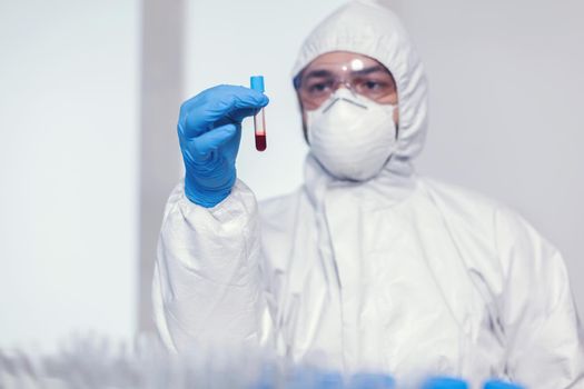 Man in coverall suit in microbiology laboratory holding test tube with blood infected with coronavirus. Doctor working with various bacteria and tissue, pharmaceutical research for antibiotics against covid19.